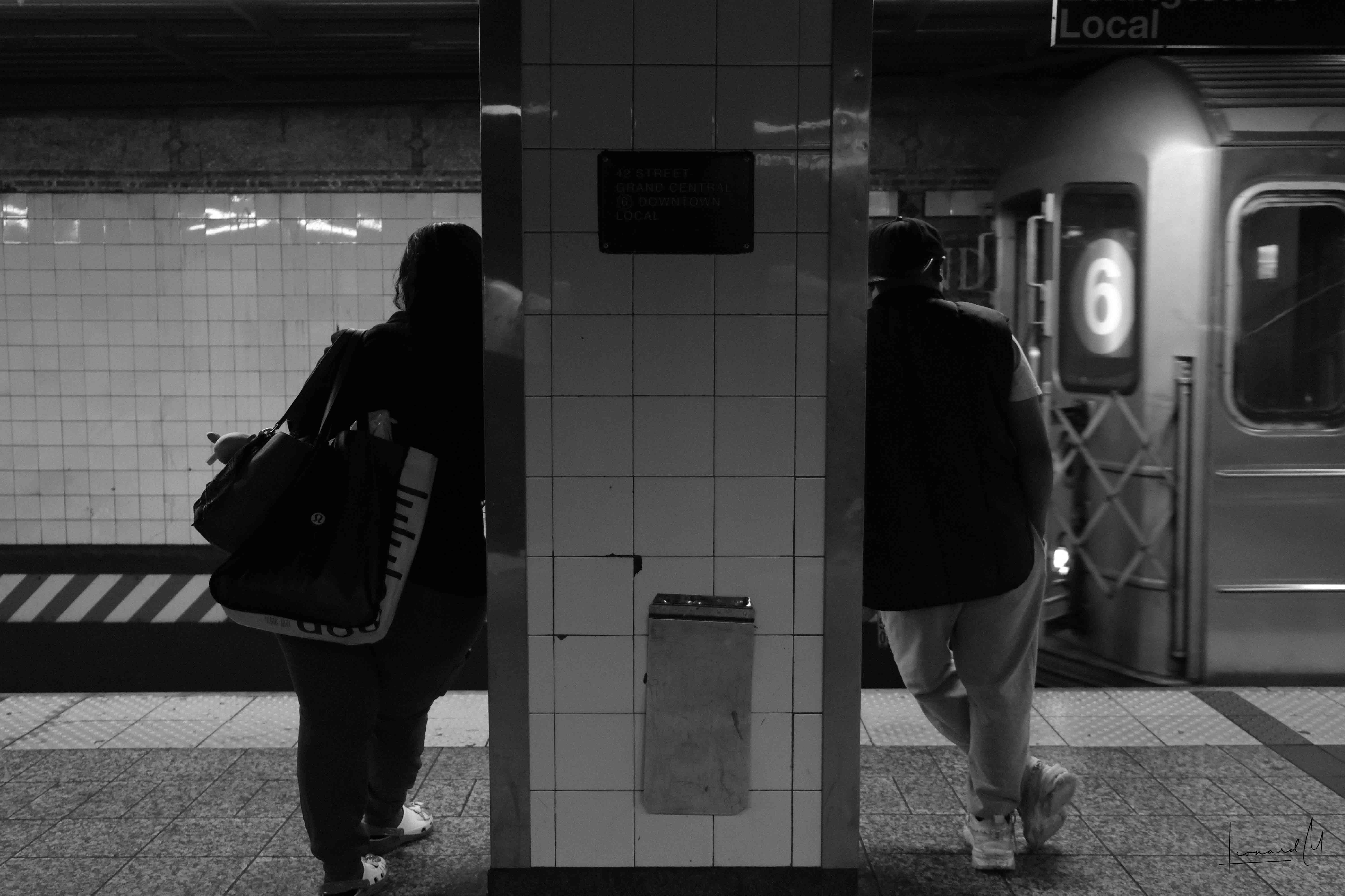 Two people leaning on a subway column from opposite sides as the 6 train arrives.